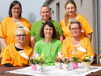 Das Team des Pflegedienstes Sonne in orangen und grünen Poloshirts am Tisch mit Frühlingsblumen